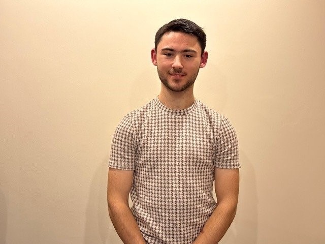 A headshot of Reece standing in front of a blank wall 