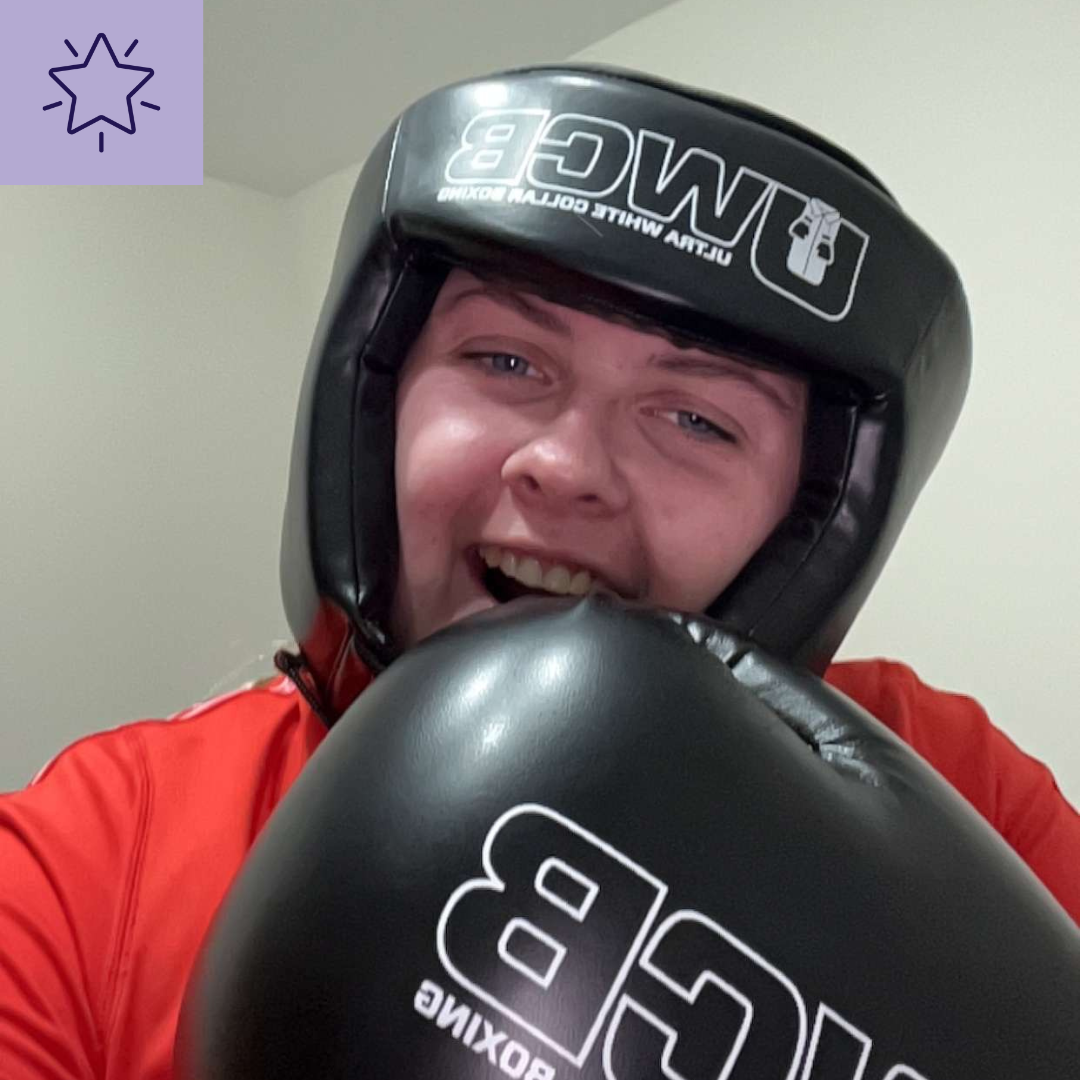 -	"Tish is smiling,  wearing a black boxing helmet with the logo 'UWCB Ultra White Collar Boxing' and matching black boxing gloves. There is a lilac, square overlay in the top left hand corner of the photo with the outline of a blue star in the middle.”